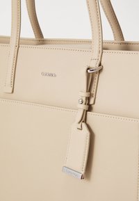 Beżowa torba typu tote marki Calvin Klein wykonana z gładkiej sztucznej skóry, z detalami przeszyć, smukłymi uchwytami oraz srebrną metką z logo i ozdobnymi elementami metalowymi.