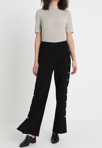 Pantalones de pierna ancha negros con acentos de botones dorados a lo largo de los lados, combinados con una camiseta de rayas de mangas cortas. Tela suave y forma acampanada.
