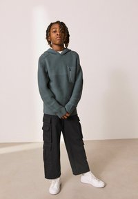 Pull en teal texturé avec capuche et poche poitrine, associé à un pantalon cargo noir avec plusieurs poches et des baskets blanches.