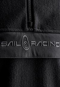 Sort fleecetrøje med halvlængde lynlås og struktureret stof. Fremtrædende "SAIL RACING" logo i en blank finish langs kanten.