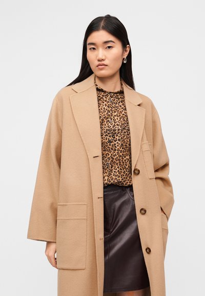 Παλτό από μαλλί καμήλας με γιακά με εγκοπές, εμπρόσθιες τσέπες και καφέ κουμπιά, φορεμένο πάνω από μπλούζα με λεοπάρ σχέδιο και δερμάτινη φούστα.