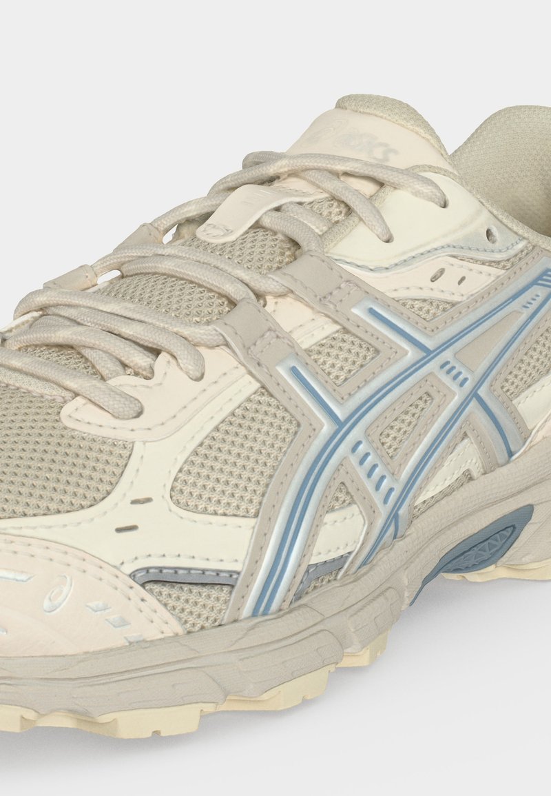 Primer plano de una zapatilla deportiva beige y azul claro con tela de malla, cordones y suela texturizada.