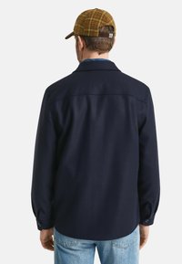 Camicia di lana blu navy con maniche lunghe, polsini con bottoni e colletto classico; abbinata a jeans blu chiaro e un cappello a scacchi.