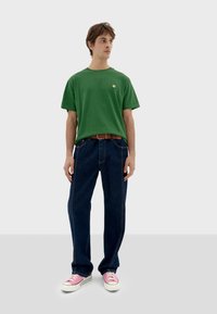 Camiseta de algodón verde con cuello redondo, combinada con jeans azul oscuro con costuras en contraste y un cinturón marrón. Llevada con zapatillas rosas.