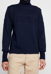 Maglione a collo alto di colore blu navy, lavorato a maglia con strisce orizzontali testurizzate, polsini a coste e tessuto morbido. Abbinato a pantaloni beige chiaro.