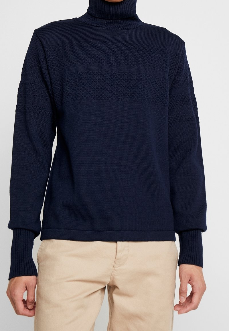 Maglione a collo alto di colore blu navy, lavorato a maglia con strisce orizzontali testurizzate, polsini a coste e tessuto morbido. Abbinato a pantaloni beige chiaro.