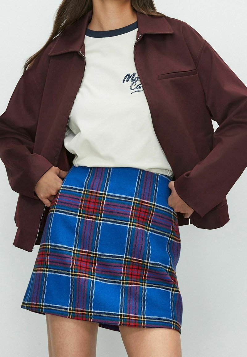 Femme portant une veste bordeaux, un t-shirt blanc à motif, et une mini-jupe à carreaux bleus, les mains reposant sur les côtés de la veste.