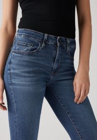 Tume sinised denimist teksad kõrge vöökohaga, viie taskuga ja õrnalt pleekinud; kontrastõmblused toovad esile õmblused ja lõppevad tõmblukuga.