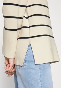 Beige gestreepte sweater met zwarte en blauwe horizontale strepen, geribbelde textuur en zijsplit, gecombineerd met lichtblauwe denim jeans.
