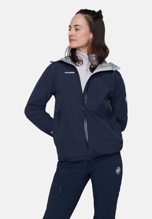 Donna con giacca e pantaloni Mammut blu navy, in piedi con le mani dietro la schiena, che guarda di lato contro uno sfondo bianco uniforme.