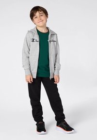 Νεαρός αγόρι στέκεται, φορώντας γκρι φούτερ με φερμουάρ, πράσινο T-shirt, μαύρο παντελόνι και μαύρα αθλητικά παπούτσια με άσπρες σόλες και πορτοκαλί λεπτομέρειες.