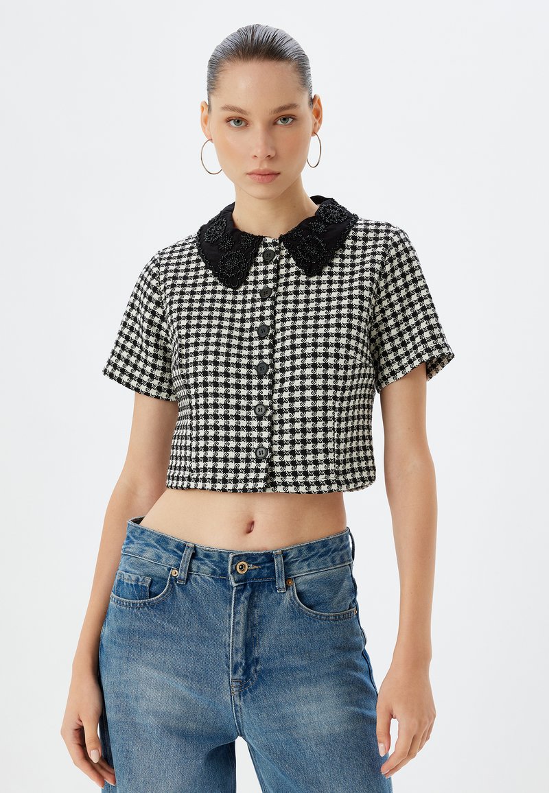 Top corto a cuadros en blanco y negro con un cuello de encaje negro decorativo, con cierre de botones y mangas cortas. Combinado con jeans azules.