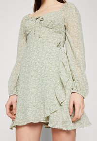 Kvinna som bär en ljusgrön, långärmad klänning med blomm önster, volanger och fyrkantig halsringning, står med händerna avslappnat vid sidorna.