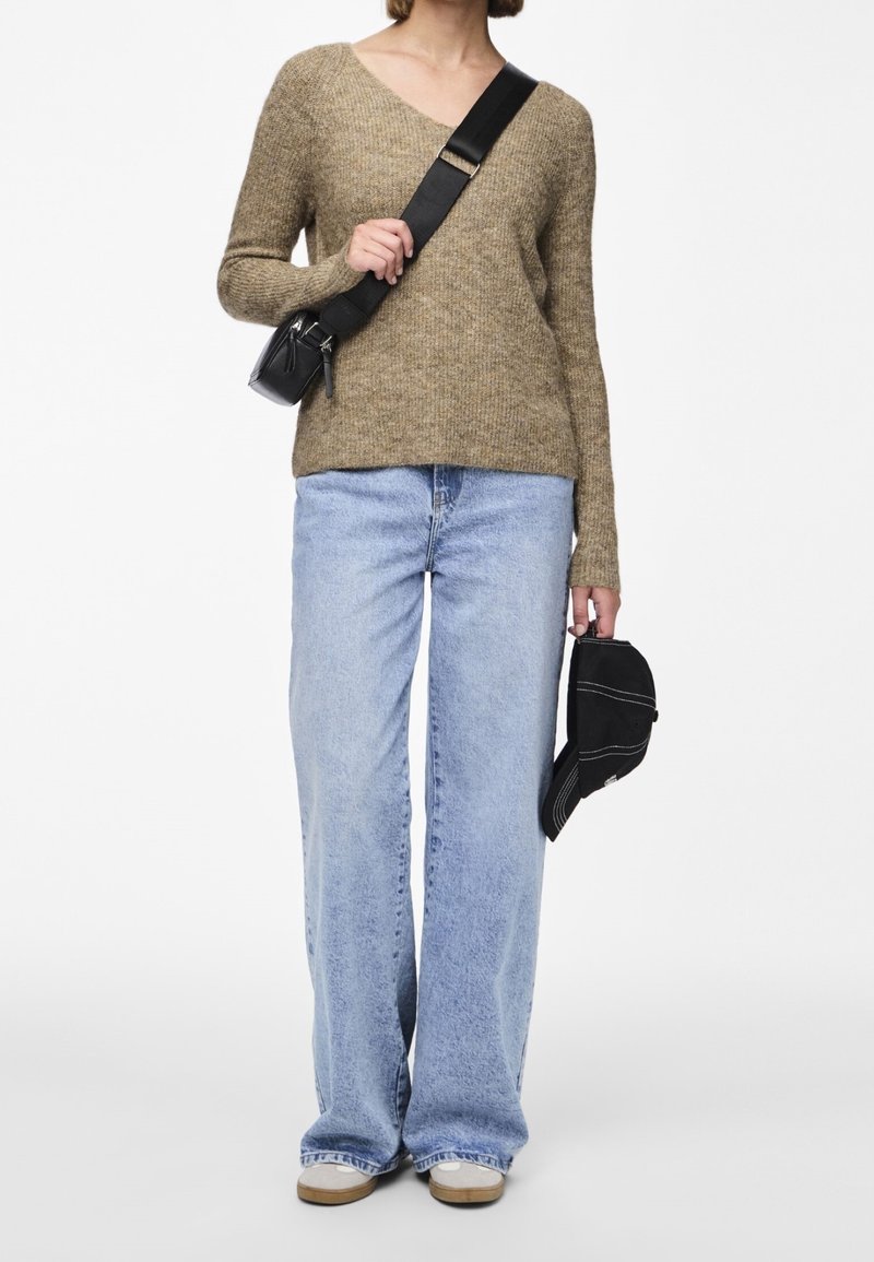 Pull marron texturé à col en V, associé à un jean bleu clair à jambes larges. Sac bandoulière noir et casquette noire tenus à la main.