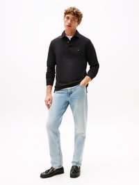 Juodas ilgomis rankovėmis polo marškiniai su apykakle. Šviesiai mėlyni džinsai. Juodi loaferiai. Modelis stovi su rankomis kišenėse prieš baltą foną.