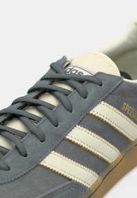 Γκρι αθλητικό παπούτσι Adidas με κρεμ ρίγες, κείμενο "Spezial" σε χρυσό και γκρι κορδόνια σε λευκό φόντο.