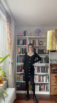 Juoda suknelė su ilgomis rankovėmis, kvadratiniu iškirpimu ir auksinėmis mygtukais. Derinama su juodais batais iki kelių. Fone matoma knygų lentyna.