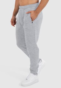 Pantalons de survêtement gris, fuselés, avec une texture lisse, une taille élastique et des poches zippées sur les côtés, associés à des baskets blanches.