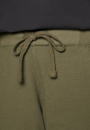 Olijfgroene joggingbroek met een gebonden koord in de taille, gedragen met een zwarte top die gedeeltelijk zichtbaar is op de taille.