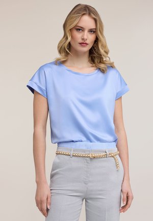 Blonde vrouw met een lichtblauw, kort mouw blouse, ingestopt in een lichtgrijze broek met een gevlochten gouden riem, staand tegen een effen achtergrond.