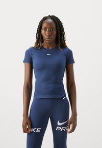 Kvinne med lange fletter iført en tettsittende marineblå Nike-skjorte og matchende leggings med synlige hvite Nike-logoer.