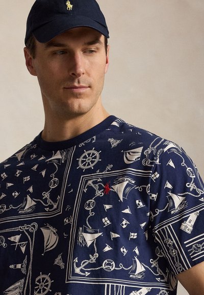 Uomo che indossa un berretto blu navy e una camicia blu navy con motivi nautici bianchi che includono ancore, timoni e barche a vela, guarda di lato.