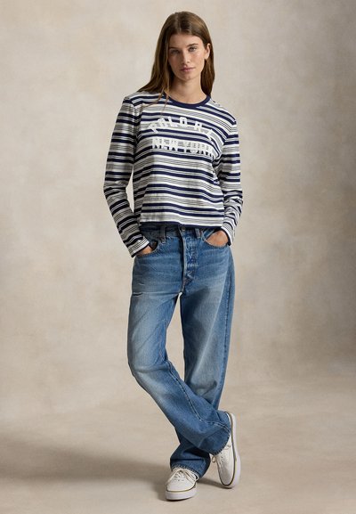 Femme portant un t-shirt à manches longues rayé bleu marine et blanc avec texte, jean bleu ample, baskets blanches, debout les mains dans les poches sur un fond neutre.