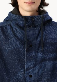 Zils rakstains jakas ar kapuci, kas izgatavota no strukturēta auduma un melniem akcentiem. Tam ir pogājama priekšpuse un regulējamas auklas.