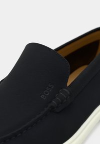 Marine slip-on sko laget av mykt materiale, med avrundet design, kontrasterende hvit såle, preget logo og subtile sømdetaljer.