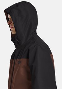 Giacca con cappuccio nera e marrone con chiusura a zip. Presenta un logo firmato sul petto e un tessuto texturizzato in design bicolore.