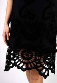 Schwarzes Kleid mit aufwendiger floralem Stickerei und einem gezackten Spitzenrand mit runden Ausschnitten, das Textur und Detail hervorhebt.