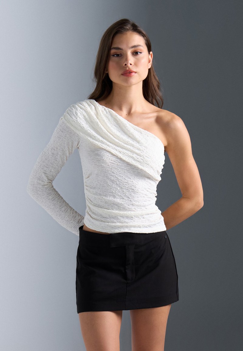 Witte one-shoulder top met textuur, gedrapeerd ontwerp en lange mouw aan één kant, gecombineerd met een zwarte minirok.