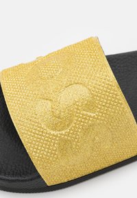 Sandale slide noire avec bride dorée texturée présentant un motif en relief embossé et une finition scintillante.