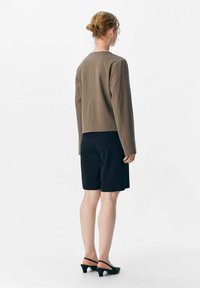 Femme avec les cheveux attachés, portant un haut taupe à manches longues, un short noir mi-cuisse et des chaussures à talons bas noires, se tenant de dos.
