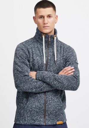Veste en polaire zippée bleu marine avec un col haut, dotée de cordons de serrage clairs contrastants, d'une fermeture éclair marron et d'un petit écusson logo sur la manche.