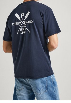 Mand iført en mørkeblå T-shirt med hvide krydsede årer og teksten "Denim Brand LDN Since 1973" på ryggen, kombineret med blå cowboybukser.