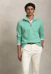 Μίντ πράσινο πουλόβερ με φερμουάρ στον λαιμό, φορεμένο πάνω από ένα ανοιχτό ροζ πουκάμισο, συνδυασμένο με κρέμ παντελόνι. Σ slim fit σχεδίαση.