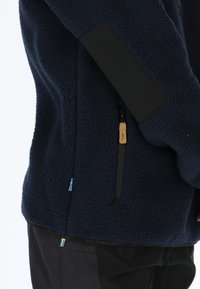 Giacca in pile navy con tessuto testurizzato, dettagli neri sulle spalle, tasca con zip e piccolo dettaglio della bandiera blu e gialla.
