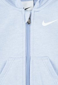 Vaaleansininen Nike-zippihiha, jossa on valkoinen swoosh-logo, etutaskut ja metallinen vetoketjun vedin, jossa on Nike-logo.