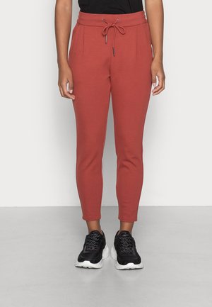 Osoba ubrana w spodnie dresowe w kolorze rdzy ze sznurkiem i czarne trampki z białymi podeszwami, stojąca na szarej podłodze przy białej ścianie.