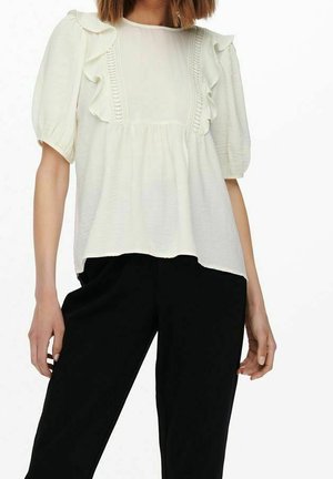 Femme portant un chemisier blanc à manches bouffantes avec des détails en volants, associé à un pantalon noir, debout devant un fond uni.