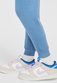 Nike sportbačiai su smėlio ir baltu viršumi, violetiniais akcentais ir šviesiai mėlynu kulnu. Suderinti su mėlynomis sportinėmis kelnėmis ir baltomis kojinėmis.