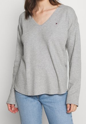 Kobieta ubrana w jasnoszary, długi rękaw koszulkę z dekoltem w serek i małym logo na piersi oraz jasnoniebieskie dżinsy, stojąca na jednolitym tle.