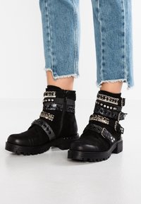 Botas negras hasta el tobillo hechas de ante, con múltiples correas texturizadas, tachuelas y un zipper lateral. Suela gruesa y diseño de puntera ligeramente redondeada.