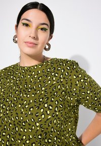Grünes Baumwollshirt mit schwarzem und gelbem Leopardenmuster, Rundhalsausschnitt und kurzen Ärmeln, getragen von einem Modell mit Creolen.