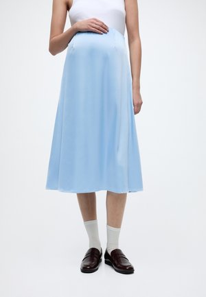 Nosečnica v beli majici brez rokavov, svetlo modri midi krilu, belih nogavicah in temno rjavih copatih, stoji na enobarvnem ozadju.