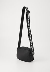 Schwarze Umhängetasche in rechteckiger Form, aus glattem Stoff, mit Reißverschluss und verstellbarem Tragegurt, der weißen Text aufweist. Minimalistisches Design.