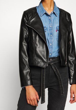 Giacca da motociclista in pelle nera con un ampio colletto, zip frontale e dettagli in cinturino borchiato, indossata sopra una camicia di denim chiaro.