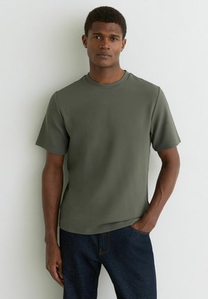 Mladý muž stojaci opretý o bielu stenu, oblečený v olivovo zelenej krátkosleeve textúrovanej tričku a tmavomodrých džínsach, s jednou rukou v vrecku.