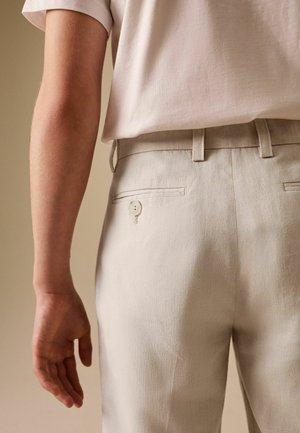 Person wearing beige trousers with back buttoned pocket and light beige short-sleeve shirt, shown from lower back to mid-arm.